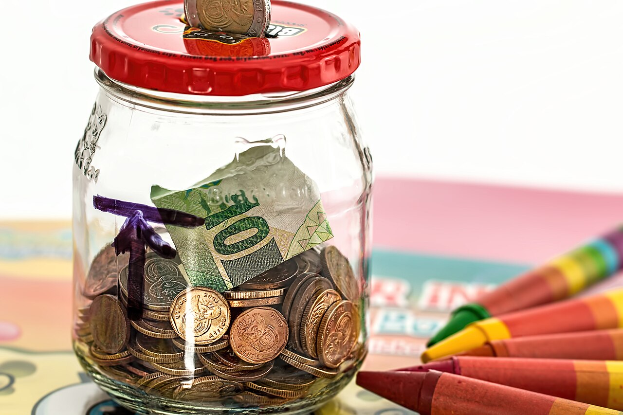 Frasco de vidro cheio de moedas de euro, com uma nota dobrada e lápis de cera ao lado.